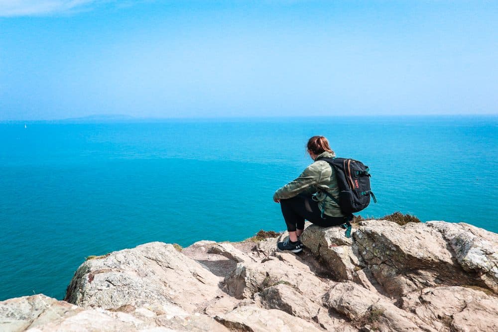 woman with a backpack on sitting on a rocky cliff looking out towards bright blue water 