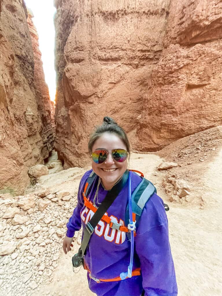 A smiling person in a Clemson University sweatshirt stands on the trail of a red rock canyon, with the towering canyon walls in the background, exuding a sense of adventure and the joy of exploration.