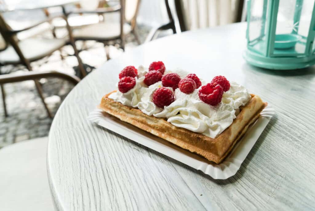 A Belgian waffle topped with whipped cream and fresh raspberries, served on a white plate on an outdoor cafe table, with chairs in the background suggesting a relaxed dining setting.