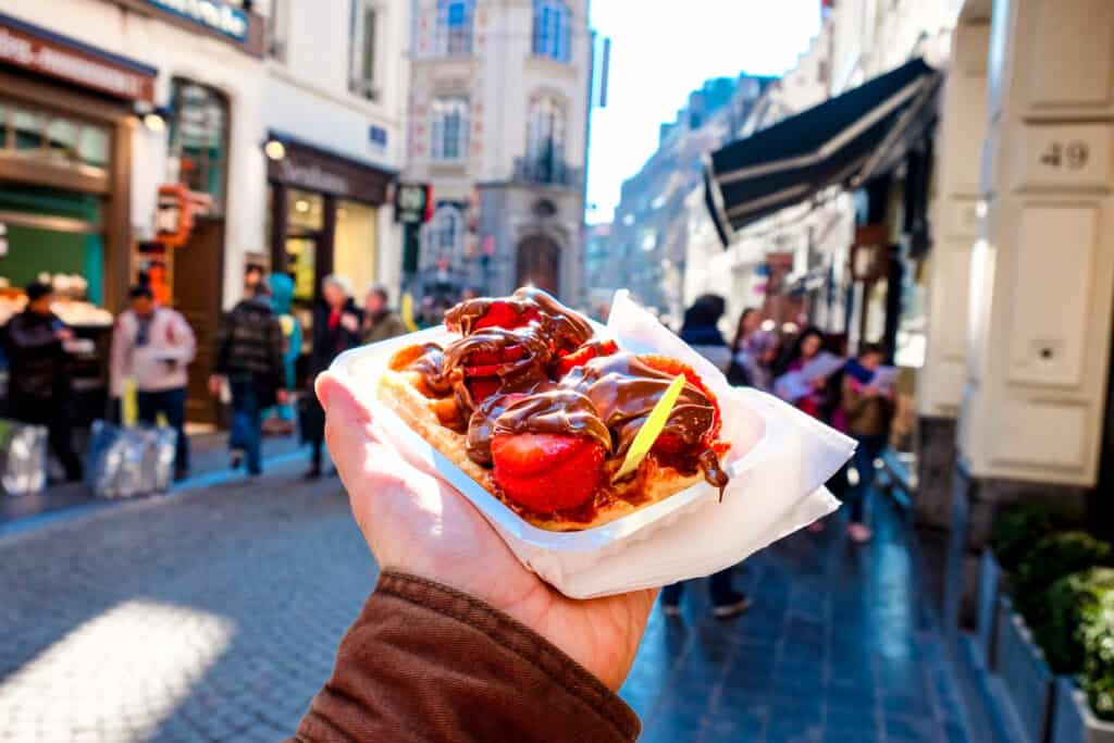 A person's hand holding a Belgian waffle topped with strawberries and drizzled with chocolate sauce, with a blurred street scene in the background capturing the ambience of a busy European city.