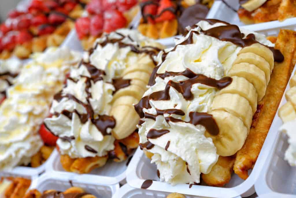 A selection of Belgian waffles topped with whipped cream, sliced bananas, and a chocolate drizzle, presented in a take-out container, ready to be enjoyed on the go.