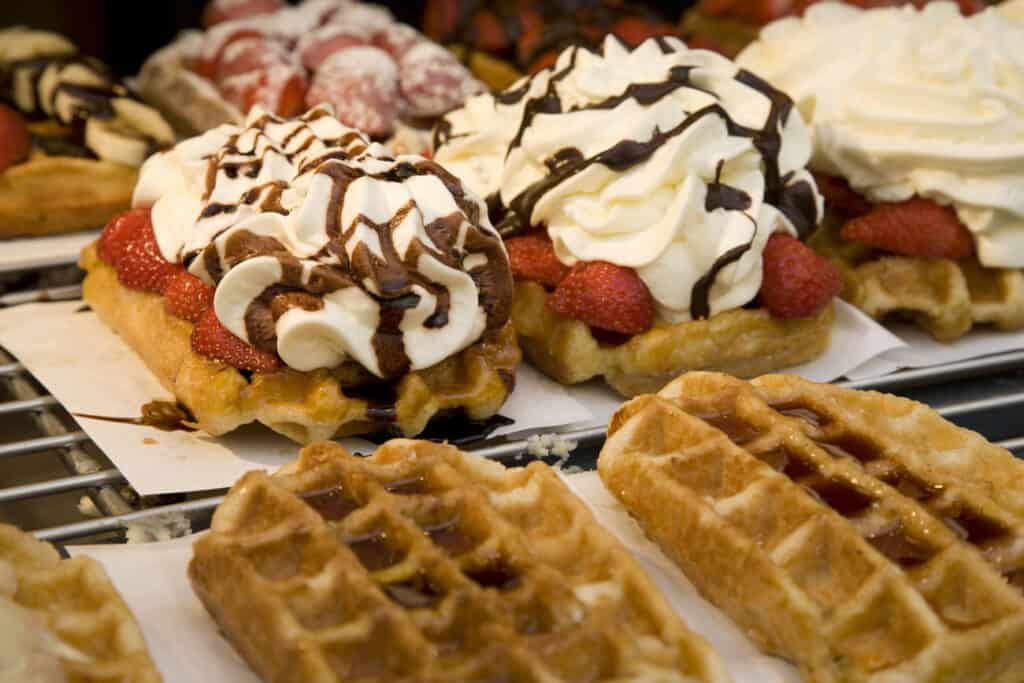 Close-up of freshly made Belgian waffles topped with whipped cream, strawberries, and chocolate drizzle, arranged on a bakery display.
