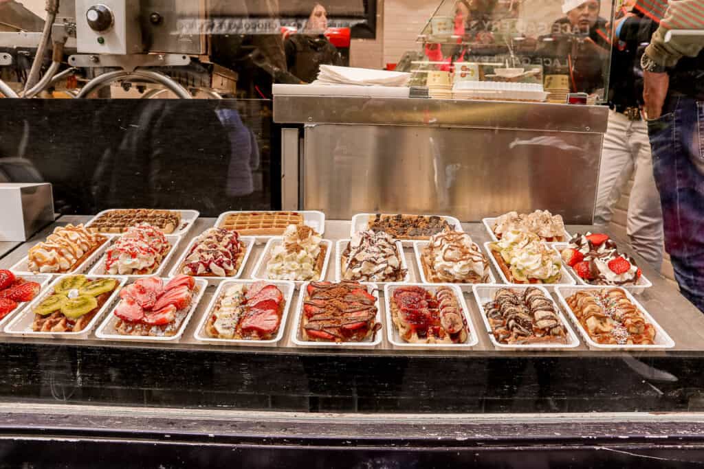 An array of Belgian waffles topped with various fruits, creams, and sauces, displayed in a street food stall with a visible waffle iron in the background, reflecting the image of onlookers.