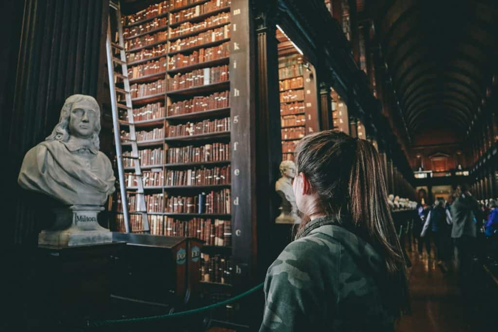 woman looking towards massive bookshelves with small statues at the end of each section 