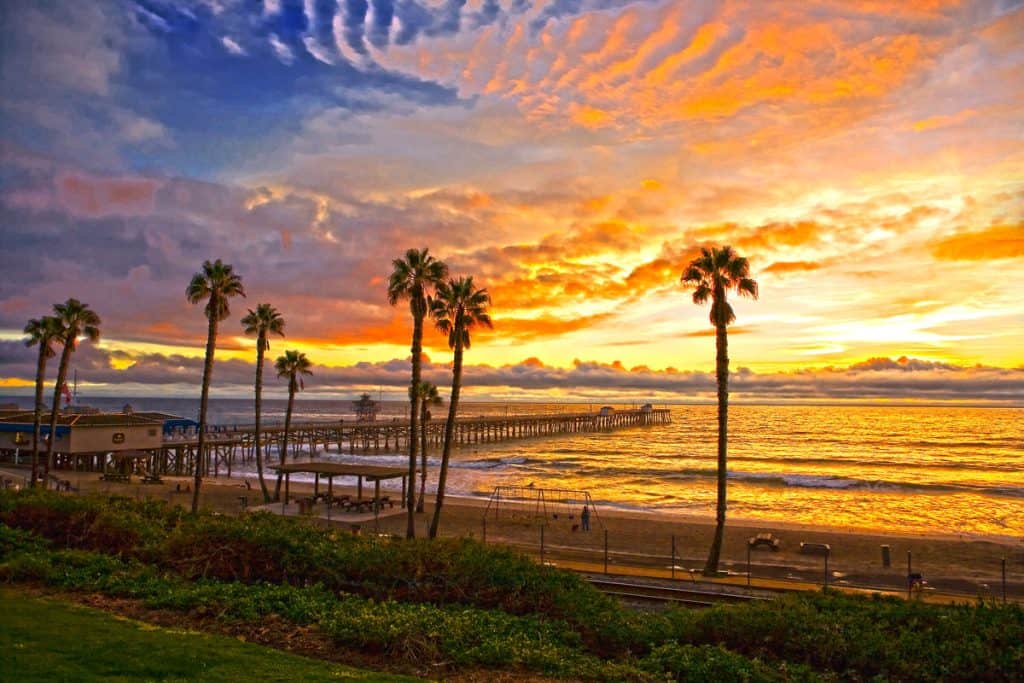 sunset at a beach with a playground, palm trees, and a long pier