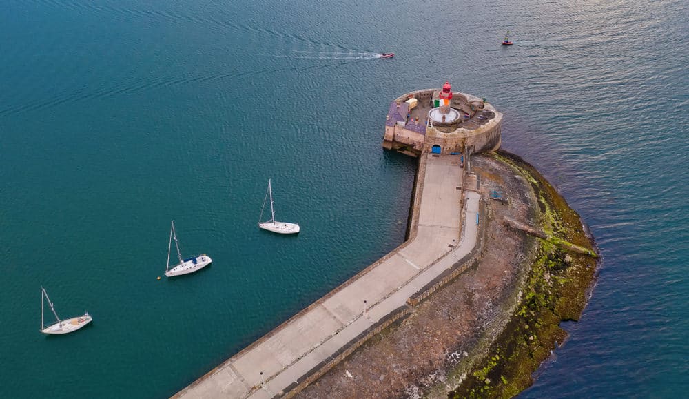 aerial view of a small lighthouse at the end of a pier with boats floating in the water around it 