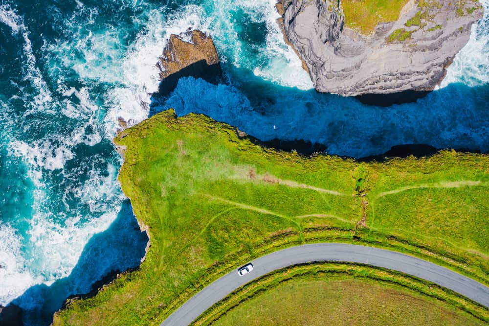 aerial view of a car driving around the edge of a grassy cliff that overlooks bright blue water 
