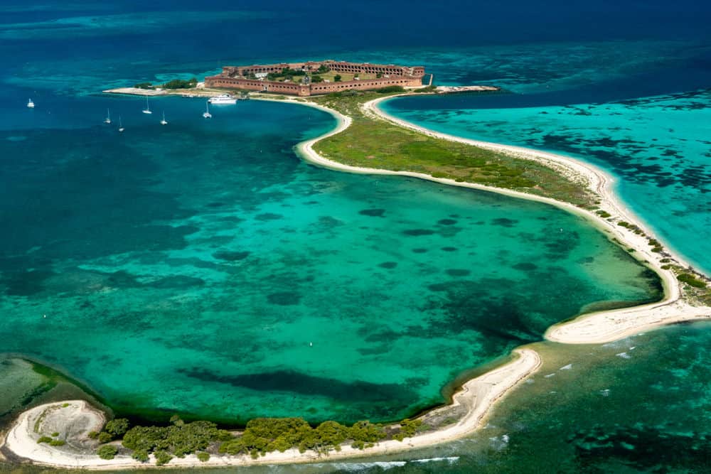 Aerial view of a sandy island with a fort surrounded by teal ocean water.
