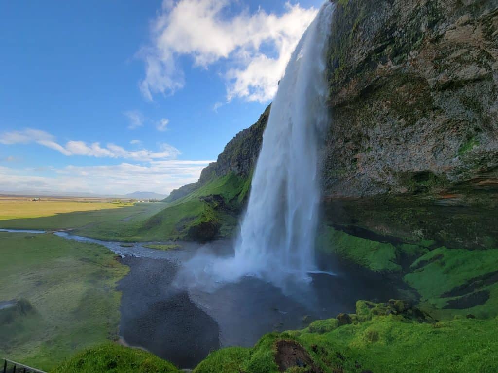 The Seljalandsfoss waterfall in Iceland rushing over a rock into a lake that flows through a grassy field. 