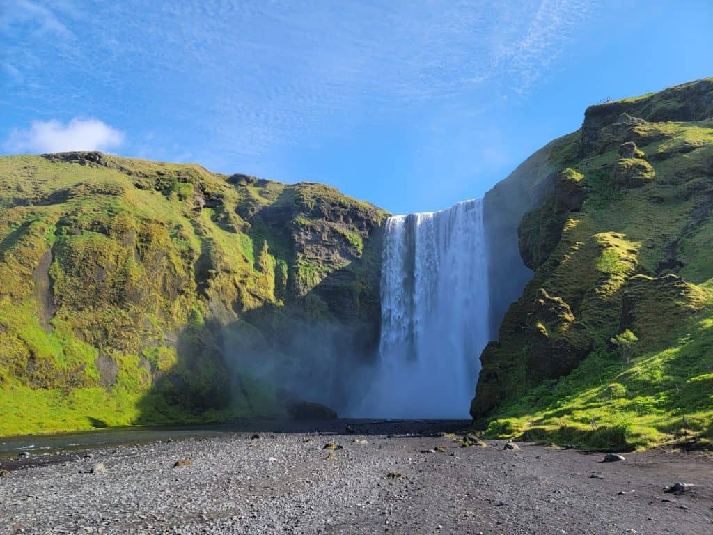 Skógafoss, one of Iceland's biggest waterfalls, rushing over the side of a cliff covered in green plants with a gravel area in front of it. 