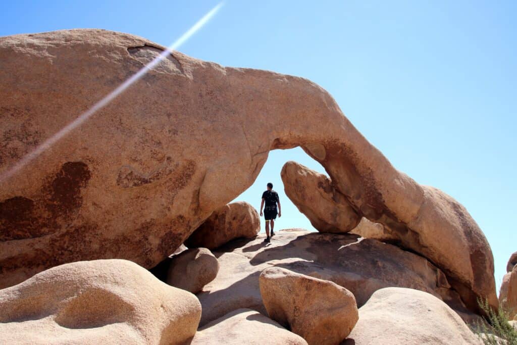 A man walking along a large rock through another rock that is arched over it. 