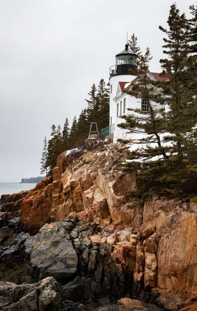 A white lighthouse perched on a rocky cliff surrounded by evergreen trees in Acadia National Park, with the ocean in the background