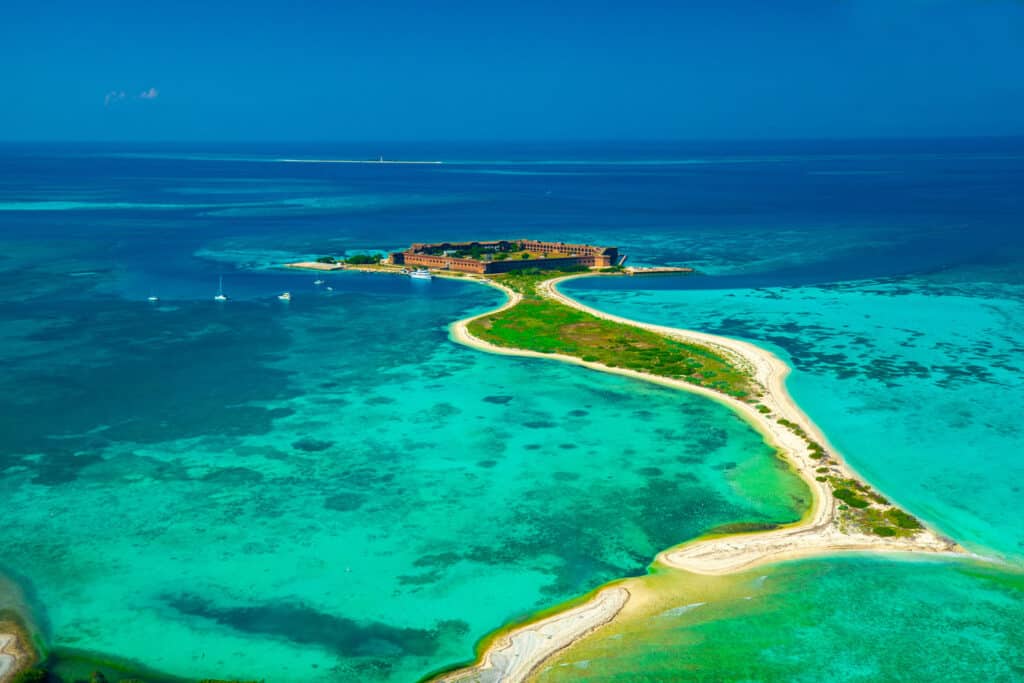 Aerial view of a narrow sandy path leading to a small island with structures, surrounded by crystal-clear turquoise waters.