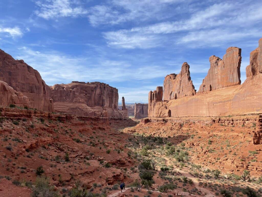 Red rocky cliff surrounded by red clay dirt and 2 hikers walking through the scenery