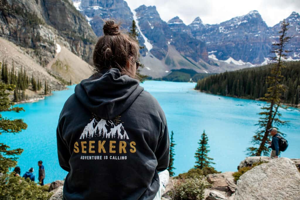 Rear view of hiker in sweatshirt with text that reads seekers adventure is calling overlooking bright blue water with snowy mountains in background