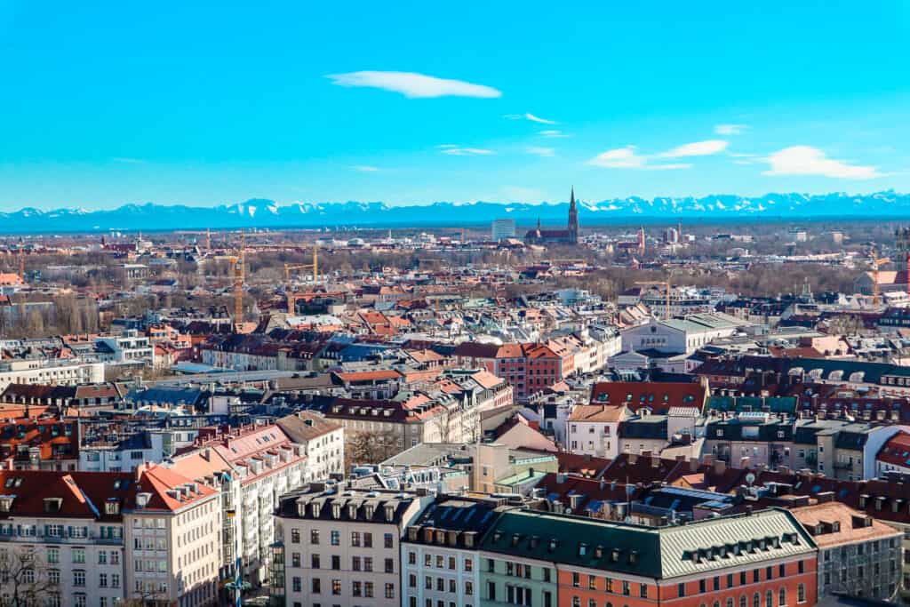 A panoramic view of Munich's cityscape with distinct European architecture, the prominent spire of the New Town Hall in the distance, and the snow-capped peaks of the Alps on the horizon under a clear blue sky.