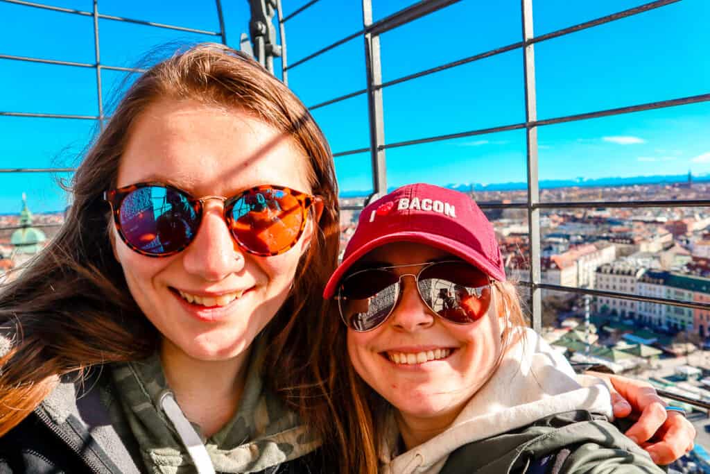 Two smiling women taking a selfie with the Munich cityscape in the background; one is wearing sunglasses and a maroon cap with "BACON" written on it, and they're both framed against a grid fence with the city below.