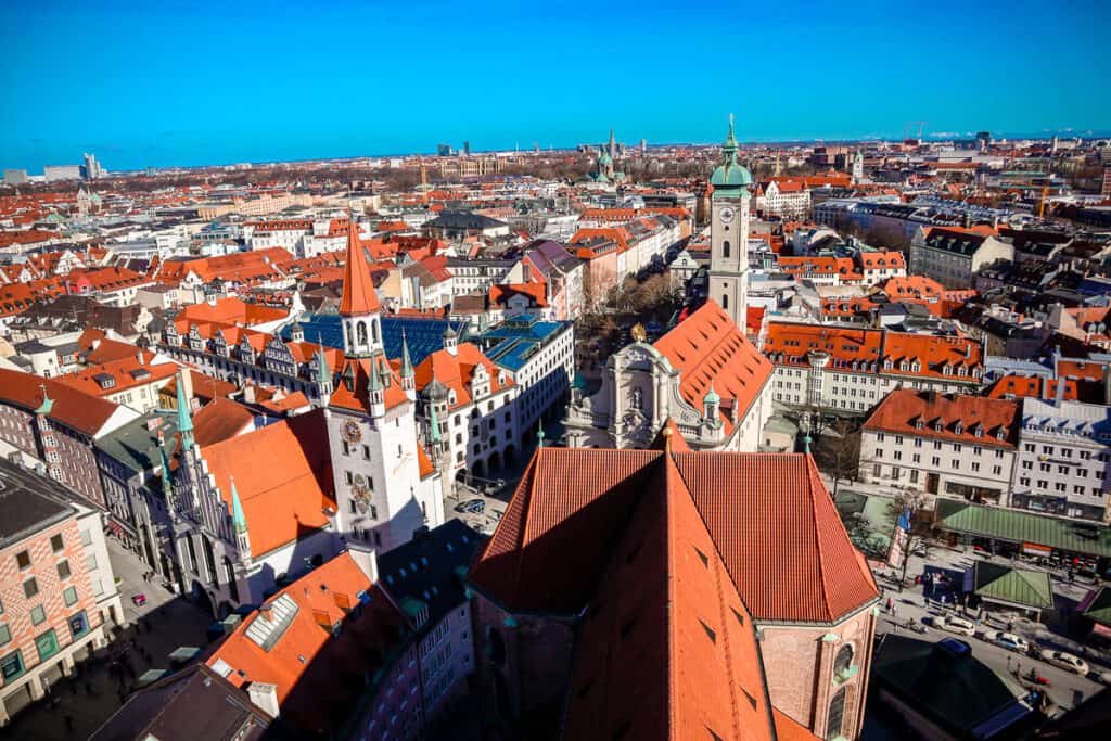 An aerial view of Munich featuring the red roofs and historical buildings of the Old Town, with the twin towers of the Munich Cathedral (Frauenkirche) and the clear blue sky above.