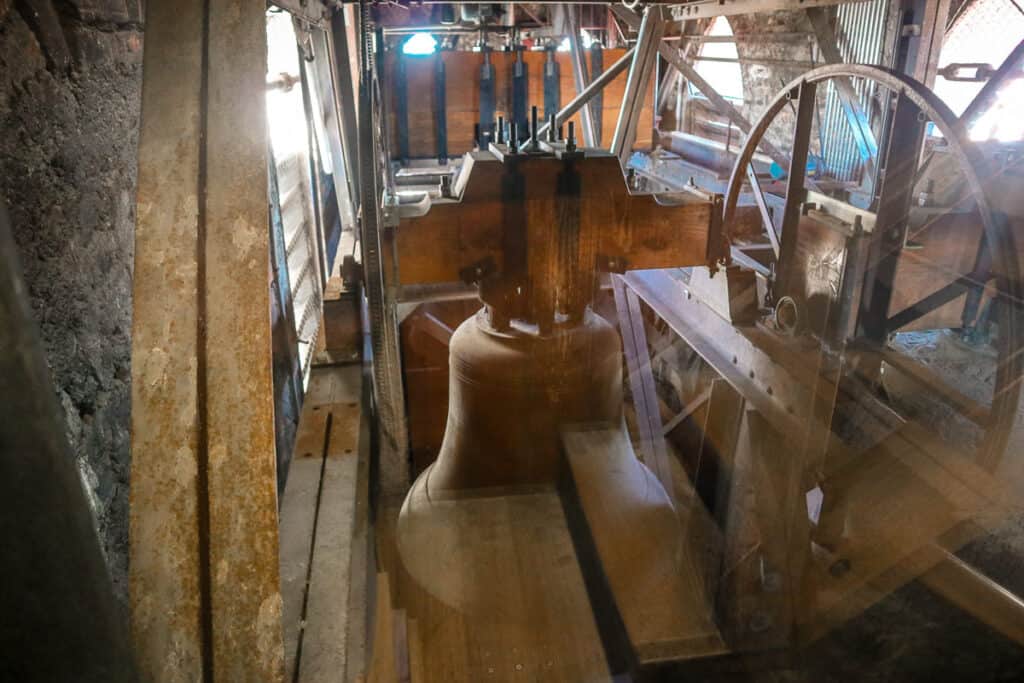 The inside of a bell tower with a large bronze bell, wooden beams, and metal mechanisms, illuminated by natural light coming through a small window.