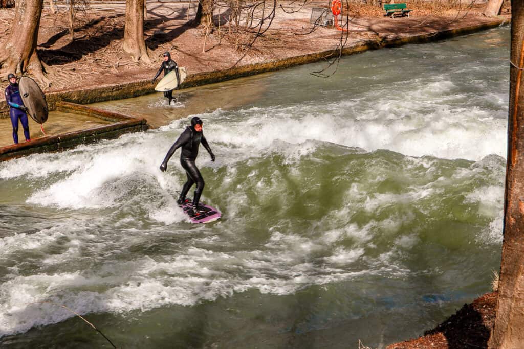 An urban surfer rides a wave in a river channel, with observers holding surfboards on the embankment, showcasing Munich's unique river surfing culture.