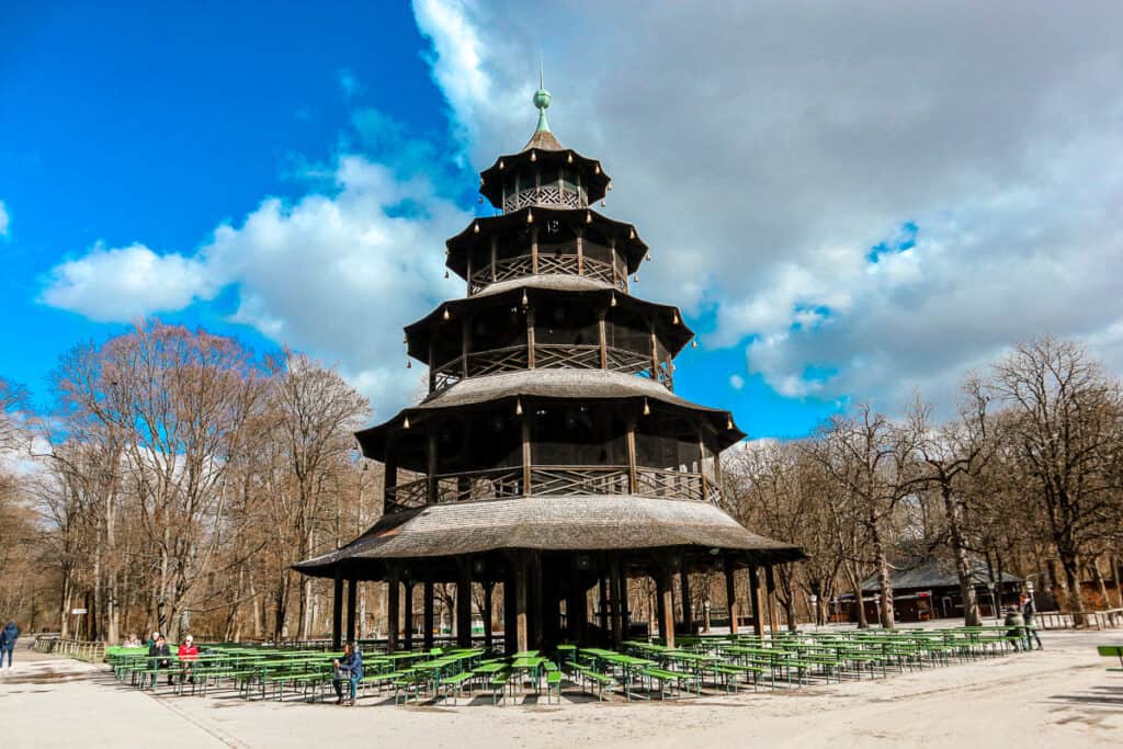A traditional wooden Chinesischer Turm (Chinese Tower) stands in the Englischer Garten in Munich, with surrounding empty beer garden benches under a partly cloudy sky.