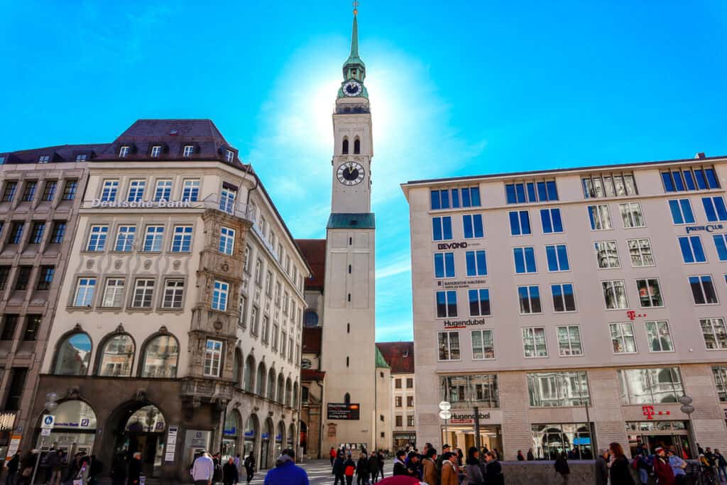 Pedestrians bustling around the Marienplatz with the iconic Glockenspiel Tower of the New Town Hall in Munich rising against a bright blue sky.