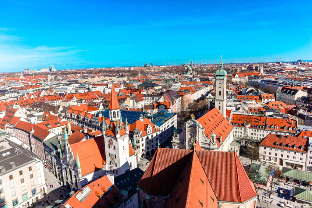 An aerial view of Munich's cityscape showing the clustered red roofs, church spires, and the sprawling urban layout under a clear sky.