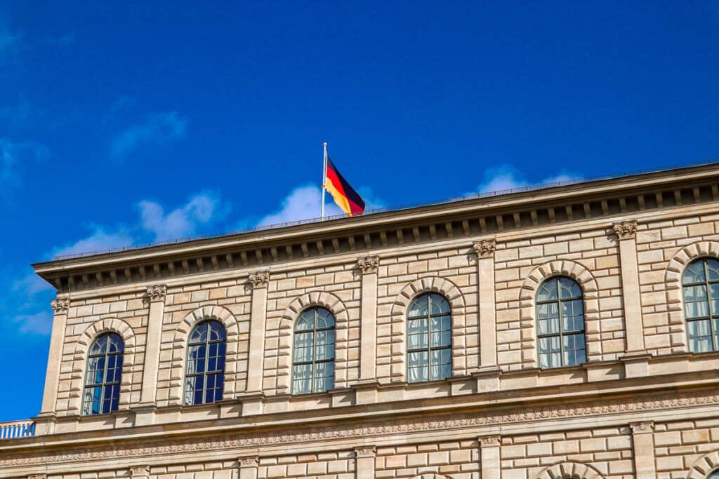 A vibrant German flag flutters against a clear blue sky atop the ornate façade of a neoclassical building with arched windows.