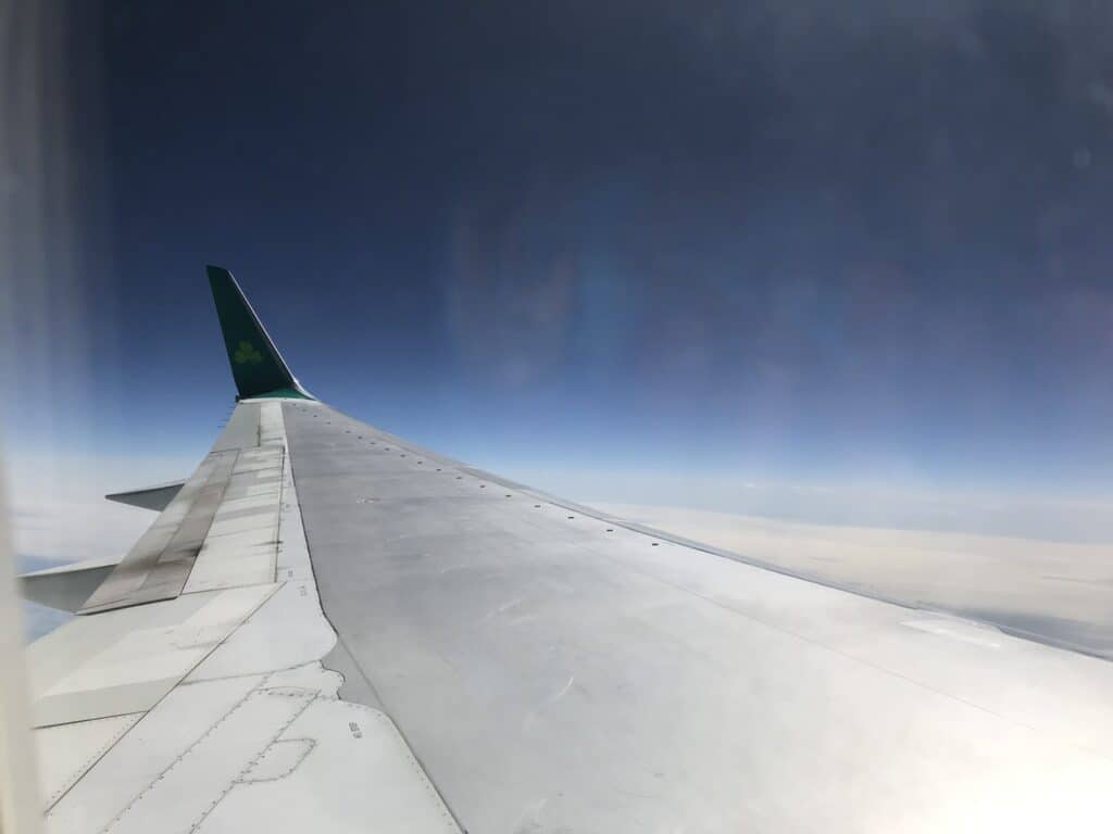 A view from the window seat of an airplane showing the wing of the aircraft with the Aer Lingus shamrock logo, flying high above the clouds.