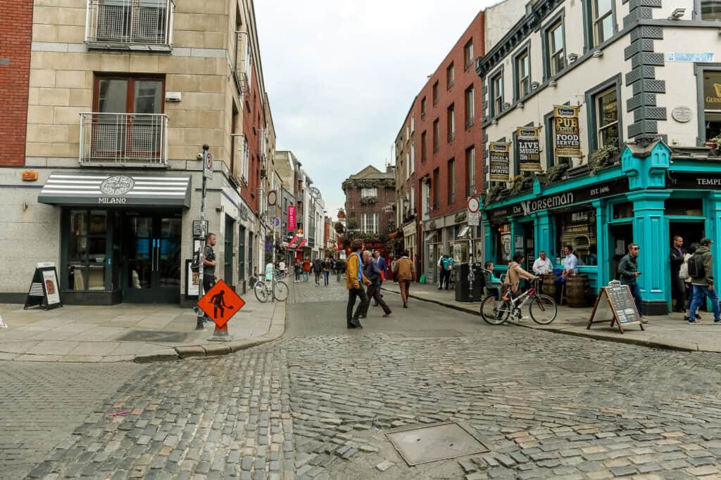 A bustling Dublin street scene featuring a mix of modern and traditional buildings, with pedestrians and cyclists navigating around a roadwork sign.