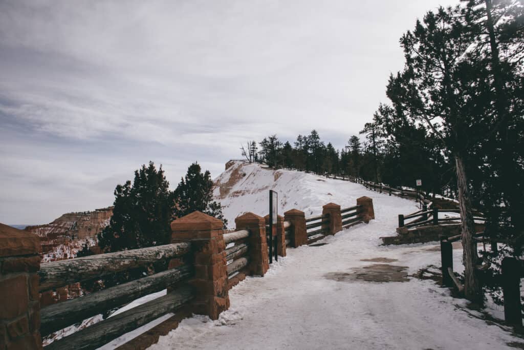 A snowy pathway leads to a viewpoint at Bryce Canyon National Park, bordered by rustic wooden fences and stone pillars. Patches of snow cover the ground and coniferous trees line the walkway, with a backdrop of distant red rock formations under a cloudy winter sky.