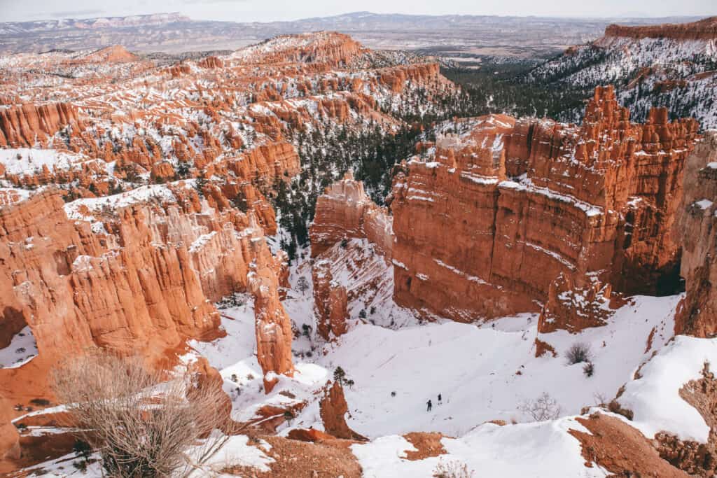 Overlooking Bryce Canyon's iconic hoodoos and natural amphitheaters, dusted with snow, highlighting the striking contrast between the vivid red rock formations and the white snow, with hikers visible in the distance, underscoring the grand scale of the landscape.