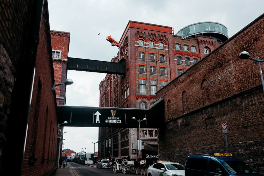 The historic red-brick architecture of the Guinness Storehouse in Dublin, connected by high walkways, with a sign pointing towards the entrance, under an overcast sky.