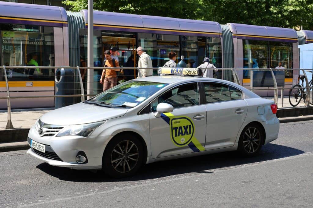 A white taxi cab in Dublin, parked by the curb in front of a tram, ready to take passengers to their destination.